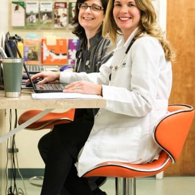 Team members smiling at camera in lobby of clinic. on left has brown hair and glasses, woman on right has blonde hair and white coat